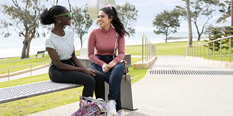Two female students sitting on a park bench, looking at each other and smiling
